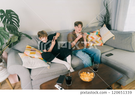 Two teenager boys are focused on their video game consoles while sitting on couch at home. Brothers Gamers enjoying Playing Video Games indoors. Siblings Spending time at home with snacks and drinks. 120651339