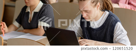 Website header image of young dreadlocked male student studying onlaptop during lecture class at university. Concentrated young man with tattoo on arm writing on computer 120651520