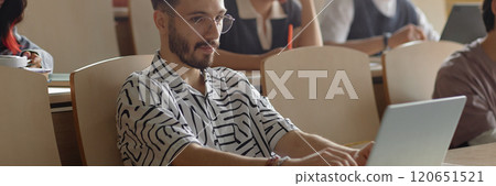 Website header shot of young Middle Eastern male student with neat beard wearing glasses studying using laptop in lecture class at university, copy space 120651521