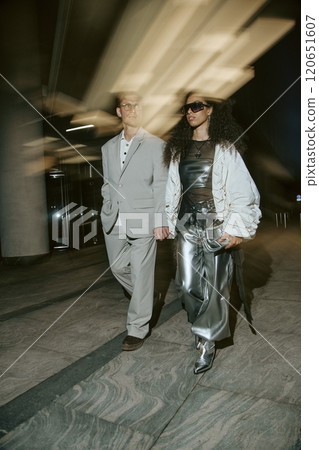 Vertical motion shot of Middle Eastern woman in sunglasses wearing silver pants holding hands with mature man in grey suit while walking in city street with in night lights in background, copy space 120651607