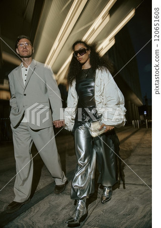 Vertical motion shot of Middle Eastern woman wearing silver trousers holding hands with mature man in classy gray suit, while walking in city street with in night lights in backdrop 120651608
