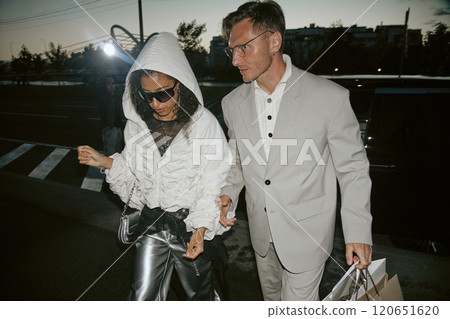 Medium shot of fashionable male celebrity wearing classy gray suit walking with female partner in trendy clothes from shopping caught by paparazzis camera lights at city street, camera flash 120651620