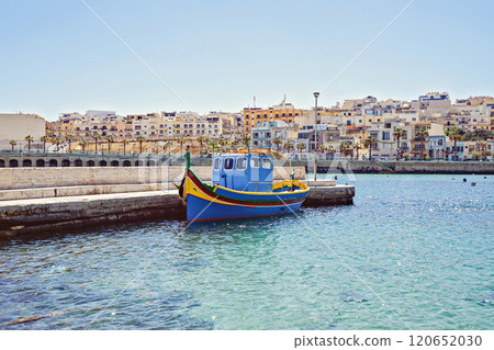 Colorful traditional Maltese fishing boat docked in calm harbor with backdrop of Mediterranean-style buildings under clear blue sky 120652030