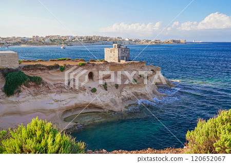 Coastal cliff with historic watch tower tower overlooking clear blue sea, lush greenery, and distant coastal town 120652067