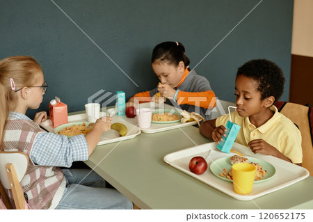 Three children of different ethnic backgrounds sitting at table together eating lunch and drinking fresh juice from carton with straw in school cafeteria, copy space Three children of different ethnic backgrounds sitting at table together eating lunch and drinking fresh juice from carton with straw in school cafeteria, copy space 120652175