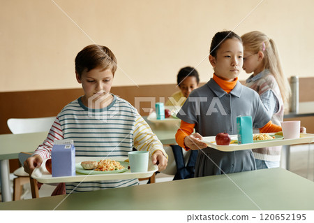 Medium shot of young boy and Central Asian girl on break, holding trays of lunch getting ready to sit down and eat at dining table in school cafeteria Medium shot of young boy and Central Asian girl on break, holding trays of lunch getting ready to sit down and eat at dining table in school cafeteria 120652195