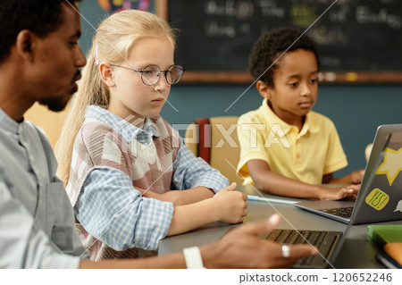 Side view of young blonde girl sitting at desk with laptop listening attentively to teachers instructions on how to complete homework assignment using laptop in modern classroom, copy space 120652246