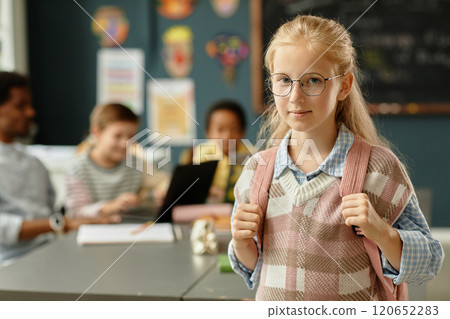 Portrait of adorable young schoolgirl with backpack wearing glasses posing in classroom and looking at camera with slight smile, copy space Portrait of adorable young schoolgirl with backpack wearing glasses posing in classroom and looking at camera with slight smile, copy space 120652283