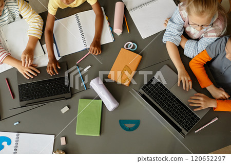 Top view of unrecognizable primary school children working in pairs using laptops to study in class while sitting at large table cluttered with stationery Top view of unrecognizable primary school children working in pairs using laptops to study in class while sitting at large table cluttered with stationery 120652297
