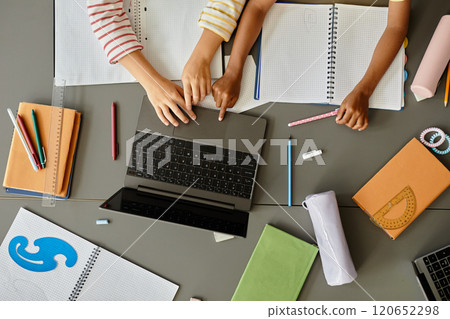 Top view of two unrecognizable young students using laptop together keeping fingers on touchpad while studying at large table cluttered with stationery Top view of two unrecognizable young students using laptop together keeping fingers on touchpad while studying at large table cluttered with stationery 120652298