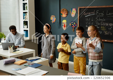 Adorable young students from diverse ethnic backgrounds standing in front of class presenting results of collaborative task to classmates during lesson in modern classroom 120652306