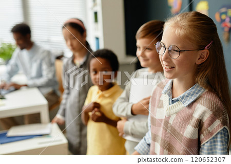 Side view of smiling young girl wearing glasses standing in line with classmates talking in front of class during lesson in primary school classroom, copy space 120652307