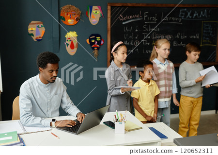 Concentrated African American male teacher typing on laptop sitting at desk, while group of young students standing at blackboard presenting homework results in classroom with blue walls Concentrated African American male teacher typing on laptop sitting at desk, while group of young students standing at blackboard presenting homework results in classroom with blue walls 120652311