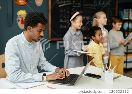 Side view of African American male teacher typing on laptop with happy children standing at classroom blackboard in background, copy space Side view of African American male teacher typing on laptop with happy children standing at classroom blackboard in background, copy space 120652312