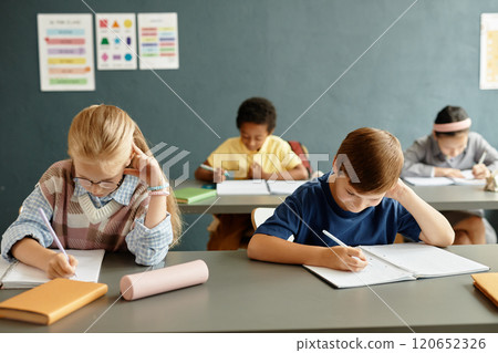 Young studious girl and boy writing in exercise books sitting at desk while studying during lesson at primary school, copy space 120652326
