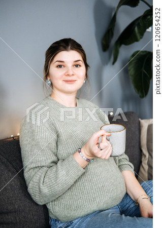 Pregnant woman enjoying a warm drink and book on cozy sofa. Autumn Winter comfort, self-care, and seasonal relaxation. 120654252