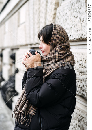 Woman in cozy scarf holding coffee cup. Urban winter fashion, layered cold-weather style, slow-living trend, sustainable fashion choices, mindful coffee moments 120654257