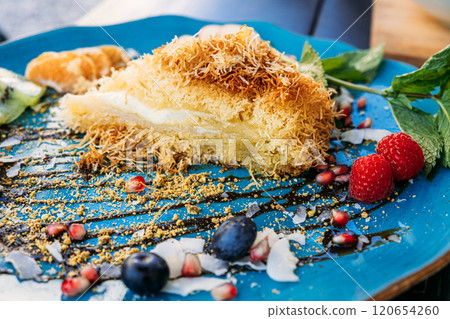 Elegant dessert with kunafa and fresh fruit slices on a blue plate. A creative presentation of Middle Eastern sweets with vibrant garnishes. 120654260