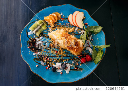Elegant dessert with kunafa and fresh fruit slices on a blue plate. A creative presentation of Middle Eastern sweets with vibrant garnishes. 120654261