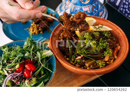 Traditional Moroccan lamb tagine served with a fresh salad. A hand scoops the dish, highlighting Mediterranean flavors and cultural culinary practices. 120654262