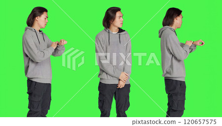 Young male model looks at watch standing against greenscreen backdrop, acting impatient in studio. Asian person feeling frustrated and irritated, waiting for a certain something. 120657575