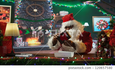 African american Saint Nick prepares a wrapped gift box in his workshop, using ribbons and bows. Santa spreading the festive holiday spirit during winter, x mas magic and fantasy. Camera B. 120657580