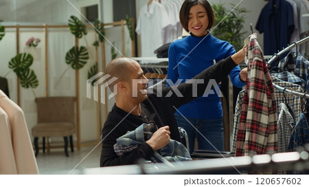 Female retail assistant taking man in wheelchair to the dressing room, helping him to try on the selection of new clothes from the racks. Asian woman ensures great customer service. Camera A. Female retail assistant taking man in wheelchair to the dressing room, helping him to try on the selection of new clothes from the racks. Asian woman ensures great customer service. Camera A. 120657602