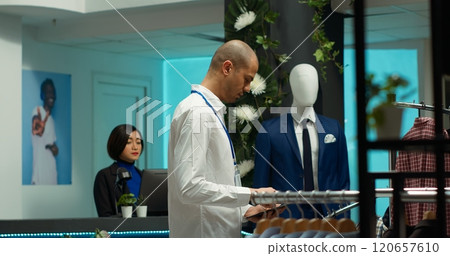 Store employee arranging new collection of clothes on display, putting merchandise on racks and emphasizing customer service. Man knows fashion knowledge, modern retail mall space. Camera A. Store employee arranging new collection of clothes on display, putting merchandise on racks and emphasizing customer service. Man knows fashion knowledge, modern retail mall space. Camera A. 120657610