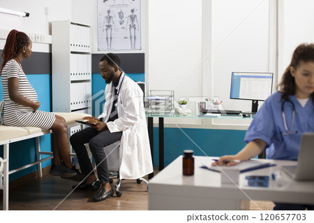 Image showcases multiethnic healthcare professionals using digital devices while pregnant woman sits on hospital bed. African american doctor discusses maternity care plan with young expectant mother. Image showcases multiethnic healthcare professionals using digital devices while pregnant woman sits on hospital bed. African american doctor discusses maternity care plan with young expectant mother. 120657703