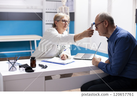 Old physician using thermometer on forehead of elderly man in hospital room, assessing his overall health. Retired female doctor checking temperature of pensioner patient during clinic appointment. 120657761