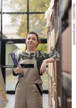 Smiling employee standing in delivery service storage, preparing clients online orders. Warehouse manager wearing industrial overall while working and coordinating goods supply chain Smiling employee standing in delivery service storage, preparing clients online orders. Warehouse manager wearing industrial overall while working and coordinating goods supply chain 120657790