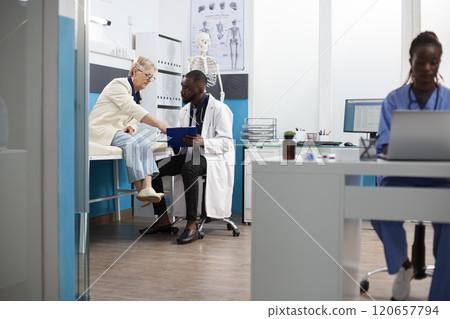 Black doctor engages in conversation with retired white woman using clipboard to outline treatment plan. Caucasian old patient asking questions as physician listens attentively in hospital room. 120657794