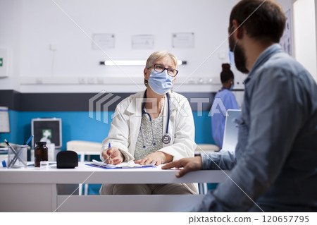 Male patient sharing sickness symptoms with friendly elderly female medic who writes on clipboard during clinic appointment. Senior doctor consults with young man in hospital, both wearing face mask. 120657795