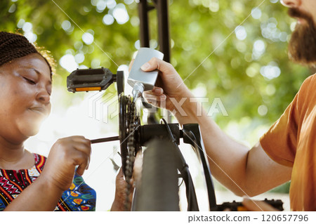 Young multiracial pair servicing and maintaining bike chain and pedals by using specialised equipment. Sports-loving boyfriend and girlfriend greasing and securing bicycle parts in home yard. 120657796