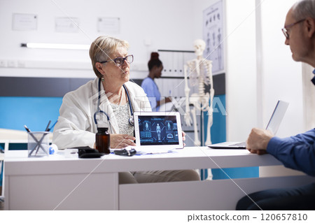 Senior woman doctor carefully guides retired male patient through diagnosis on tablet during medical checkup. Elderly physician utilizing visual aid to detail healthcare treatment options for old man. 120657810
