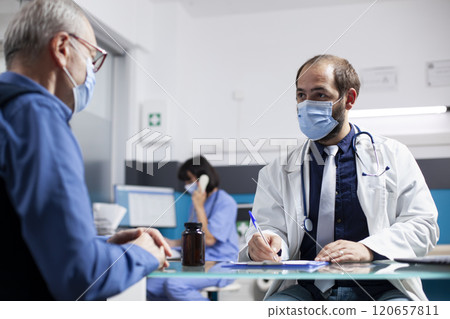 Young doctor consults with elderly male patient, attentively documenting symptoms on clipboard during medical checkup at clinic. Physician writing health concerns of retired man in hospital office. Young doctor consults with elderly male patient, attentively documenting symptoms on clipboard during medical checkup at clinic. Physician writing health concerns of retired man in hospital office. 120657811