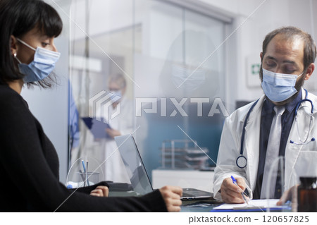 Bearded doctor writing notes for female patient during medical checkup, having plastic protective screen in between on desk. Physician reviewing health details on clipboard with sick woman in clinic. 120657825