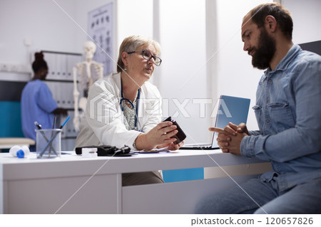 Caucasian senior doctor consults male patient, holding container of prescribed medication for recovery. Young man sits and listens to older female medical specialist give advice about his medicine. Caucasian senior doctor consults male patient, holding container of prescribed medication for recovery. Young man sits and listens to older female medical specialist give advice about his medicine. 120657826