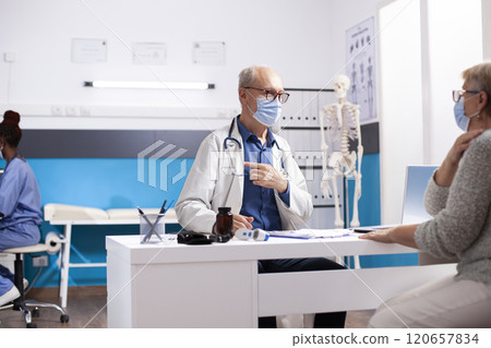 Old male doctor sits across senior white woman in clinic office, discussing checkup results, both wearing face masks for safety. Elderly medic consulting with female pensioner patient in hospital. 120657834
