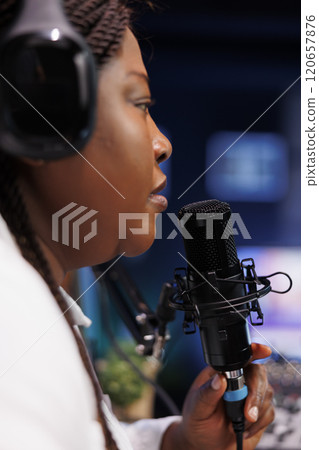 Detailed view of female influencer speaking into modern microphone, producing online talk show from home. African woman broadcasting live podcasts while utilizing wireless headphones. 120657876
