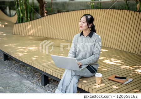 A charming Asian woman works remotely outdoors, sitting on a park bench with her laptop. A charming Asian woman works remotely outdoors, sitting on a park bench with her laptop. 120658162