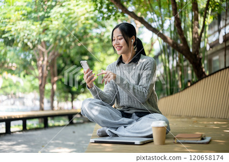 A woman sits on a park bench, selecting music on her phone, enjoys listening through her earphones. A woman sits on a park bench, selecting music on her phone, enjoys listening through her earphones. 120658174