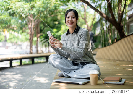 A charming Asian woman sits on a park bench, wearing earphones and smiling at the camera. A charming Asian woman sits on a park bench, wearing earphones and smiling at the camera. 120658175