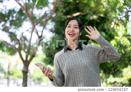 A cheerful Asian woman waves to greet her friends while walking along the sidewalk in a park. 120658197