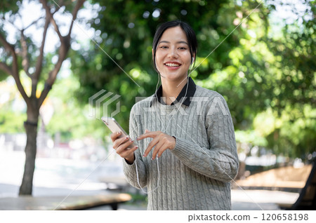 A charming Asian woman enjoys listening to music while walking along the footpath. A charming Asian woman enjoys listening to music while walking along the footpath. 120658198