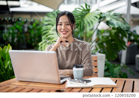 An Asian woman working remotely from a serene cafe, sitting at an outdoor table with her laptop. An Asian woman working remotely from a serene cafe, sitting at an outdoor table with her laptop. 120658200