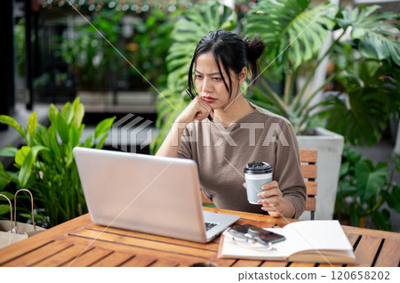 A thoughtful Asian woman looks at her laptop screen with a serious face, working remotely. A thoughtful Asian woman looks at her laptop screen with a serious face, working remotely. 120658202