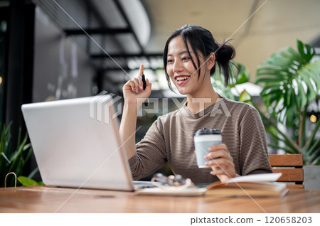 A charming Asian woman working remotely from a serene green cafe, enjoying a coffee while working. A charming Asian woman working remotely from a serene green cafe, enjoying a coffee while working. 120658203