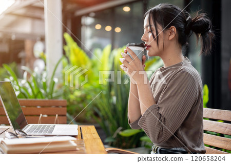A beautiful, calm Asian woman enjoys a hot drink while working remotely from a coffee shop. A beautiful, calm Asian woman enjoys a hot drink while working remotely from a coffee shop. 120658204