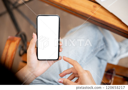 Awoman using her smartphone while sitting in a cafe. The smartphone with a white screen mockup. 120658217